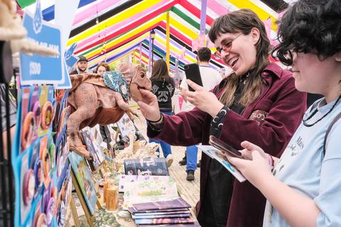 En una feria llena de coloridos stands, dos amigas se ríen mientras observan una figura de dinosaurio.