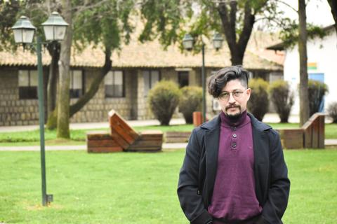 Un hombre vestido con suéter morado y abrigo negro posa en un parque con árboles y bancos de madera al fondo.