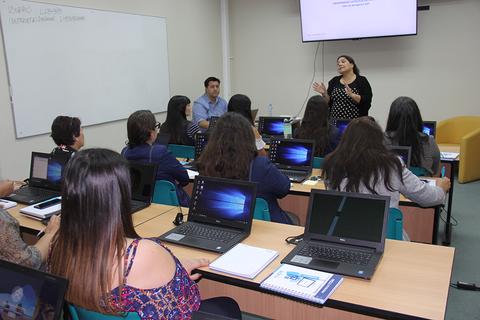 Una clase en un aula con estudiantes usando computadoras portátiles mientras una docente imparte una lección.