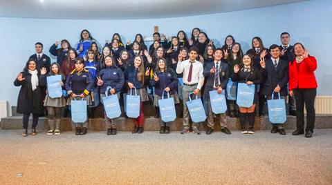 Un grupo numeroso de estudiantes sonríe y hace un gesto de saludo mientras sostienen bolsas azules.