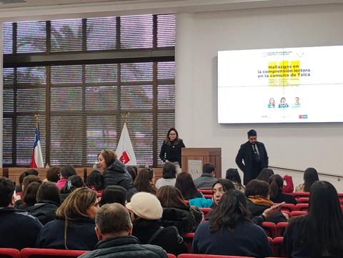 Una reunión en un salón con varias personas sentadas y dos presentadores al frente.