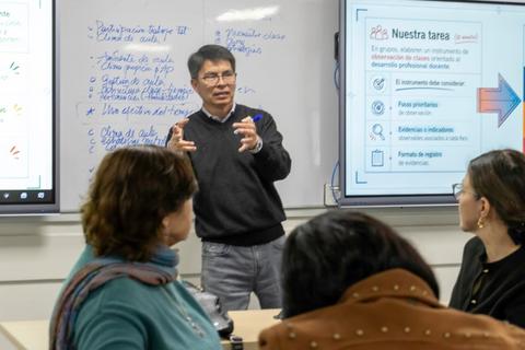 Un profesor está explicando un tema en una sala de clases mientras los estudiantes lo escuchan atentos.