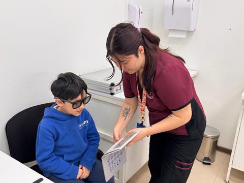 Un niño con gafas está sentado mientras una mujer le muestra una hoja en una consulta médica.