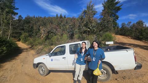 Dos mujeres sonrientes posan junto a una camioneta en un camino rural rodeado de árboles.