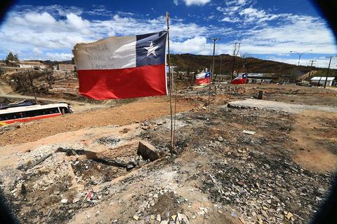 Una bandera chilena ondea en un paisaje desolado con terrenos quemados y destruidos.