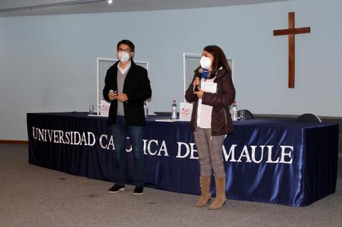 Dos personas hablan en un evento en la Universidad Católica del Maule, con un crucifijo visible en el fondo.