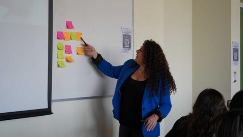 Una mujer con cabello rizado está explicando algo en una pizarra mientras señala notas adhesivas de colores.