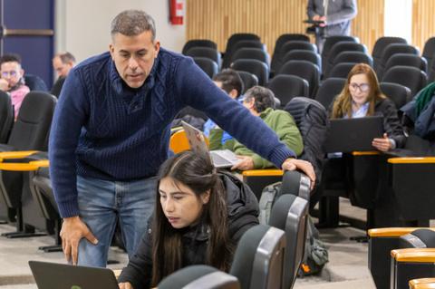 Un hombre observa a una mujer que trabaja en un portátil en un aula con asientos vacíos alrededor.