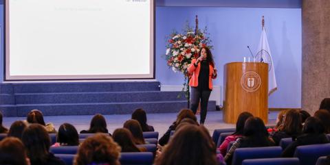 Una mujer habla frente a un auditorio lleno de personas, con un escenario decorado en el fondo.