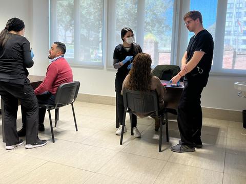 Un grupo de personas en un consultorio médico, donde dos profesionales de la salud están atendiendo a pacientes sentados en una mesa.