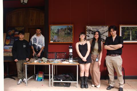 Un grupo de cinco personas posa frente a una mesa decorada con materiales, en un espacio con obras de arte en las paredes.