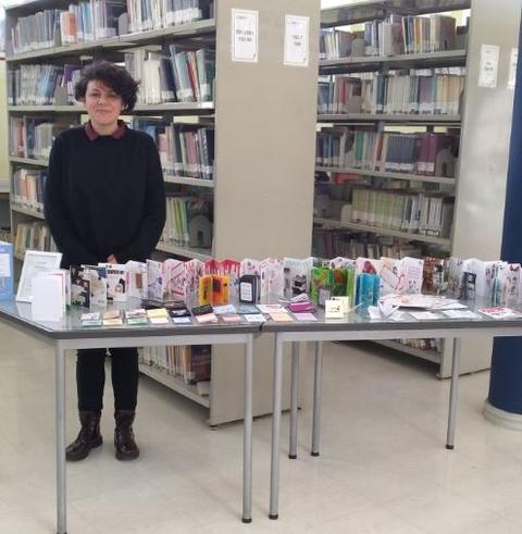 Una persona de pie junto a una mesa repleta de libros y materiales en una biblioteca.