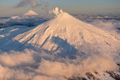 Un paisaje montañoso en el que se destacan dos volcanes cubiertos de nieve, uno de ellos emitiendo humo.
