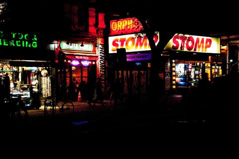 La imagen muestra un barrio iluminado por neones en una noche oscura, con distintos letreros de tiendas y restaurantes.