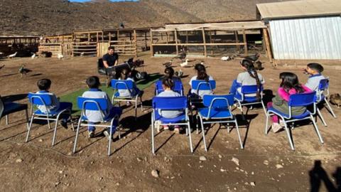 Un grupo de niñitos sentados en sillas escuchando a un profesor en un entorno rural.