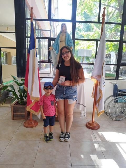 Una mujer y un niño posan juntos frente a unas banderas y una imagen religiosa en un ambiente luminoso.