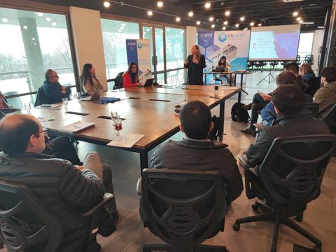 Una reunión en una sala con un grupo de personas sentadas alrededor de una mesa, mientras una mujer hace una presentación.