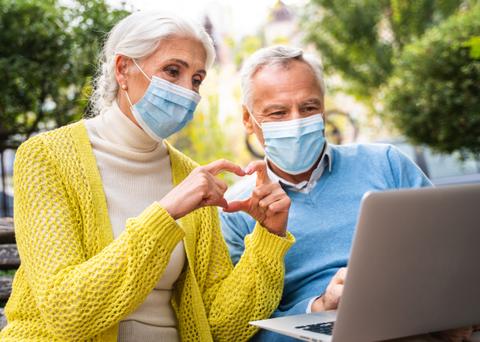 Una pareja mayor que lleva mascarillas sonríe mientras hace un gesto de corazón con las manos frente a un ordenador portátil.