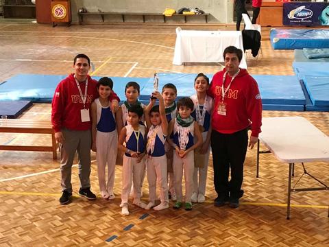 Un grupo de niños y entrenadores posan felices sosteniendo trofeos tras una competencia de gimnasia.