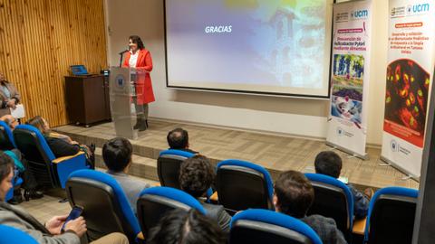 Una mujer con chaqueta roja está hablando en un escenario frente a una audiencia sentada.