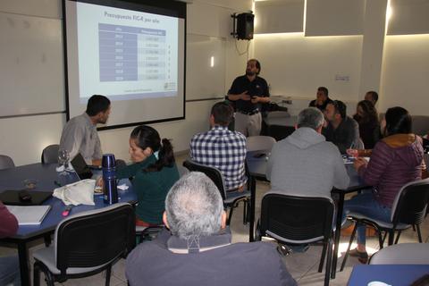 Una reunión donde un presentador expone datos a un grupo de personas sentadas alrededor de una mesa.
