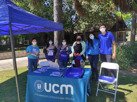 Un grupo de personas se encuentra en un stand informativo de la Universidad Católica del Maule, con un toldo y mesas en un ambiente exterior.