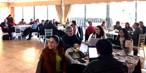 Un grupo de personas sentadas en una sala con mesas, algunas usando computadoras portátiles.
