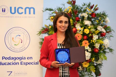 Una joven sonriente sostiene un premio frente a un fondo de flores y un banner de la universidad.