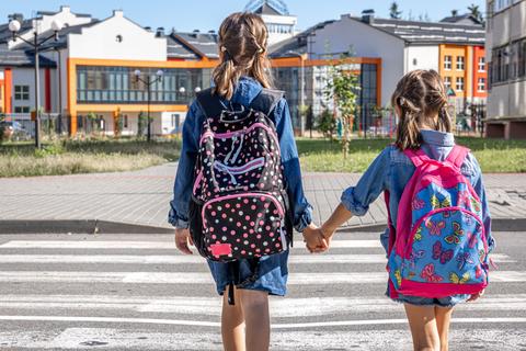 Dos niñas de la mano caminan hacia la escuela con mochilas coloridas.