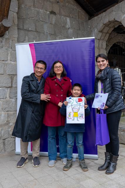 Un niño muestra orgullosamente su dibujo junto a tres adultos en un evento al aire libre.