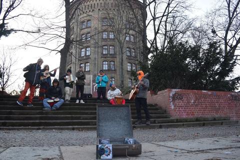 Un grupo de personas se encuentra en una escalera, con un músico tocando la guitarra y varios oyentes alrededor en un ambiente urbano.