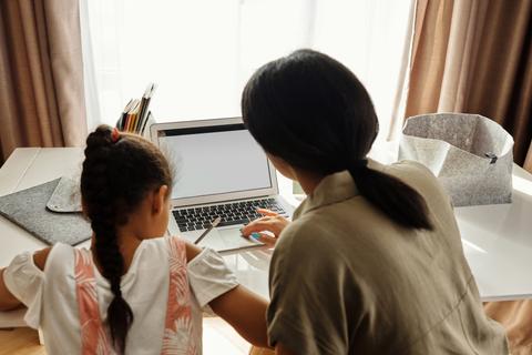 Una madre y su hija están sentadas frente a una computadora portátil en una mesa iluminada por la luz natural.