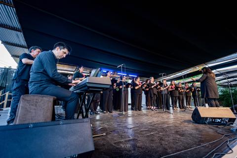 Un grupo de cantores se presenta en un escenario al aire libre, mientras un músico toca el teclado.