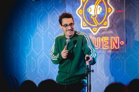 Un comediante actúa en un escenario con un micrófono, mientras sonríe y se dirige al público.
