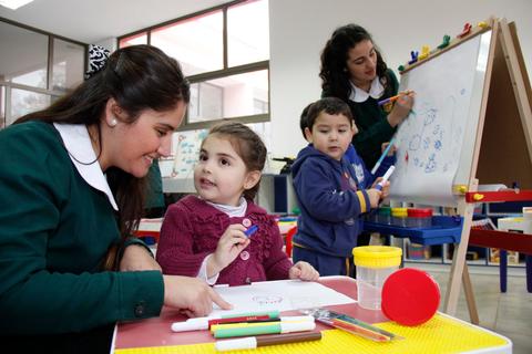 Una maestra interactúa con niños pequeños mientras dibujan en un aula colorida.