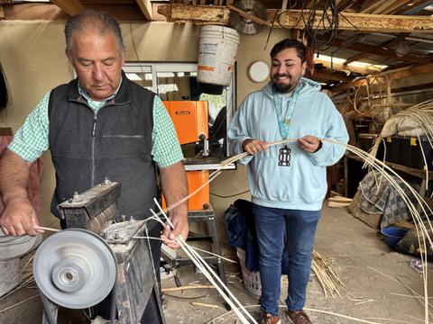 Dos hombres trabajan en un taller, uno usa una máquina para procesar cañas mientras el otro observa y sonríe.
