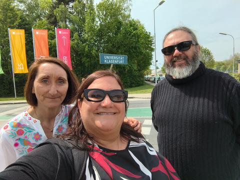 Tres amigos posando felices frente a la Universidad de Klagenfurt en un día soleado.