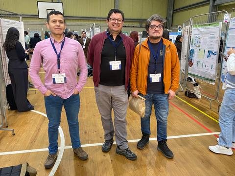 Tres hombres posando en un evento académico con trabajos de investigación detrás.