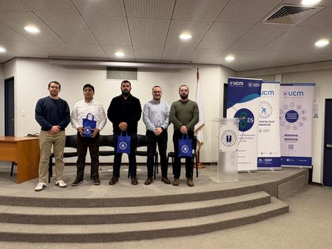 Un grupo de cinco personas se encuentra posando en un escenario con banners de la UCM al fondo.
