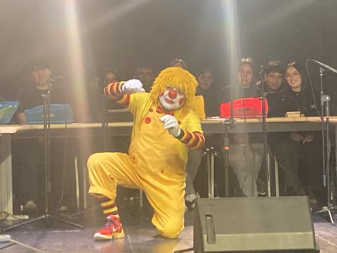 Un payaso vestido de amarillo haciendo una divertida pose en un escenario ante un público.
