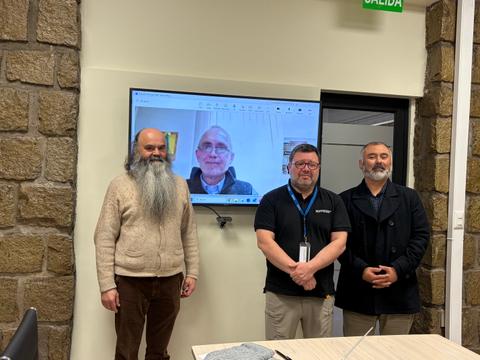 Un grupo de tres hombres posando frente a una pantalla donde aparece un cuarto hombre en videollamada.