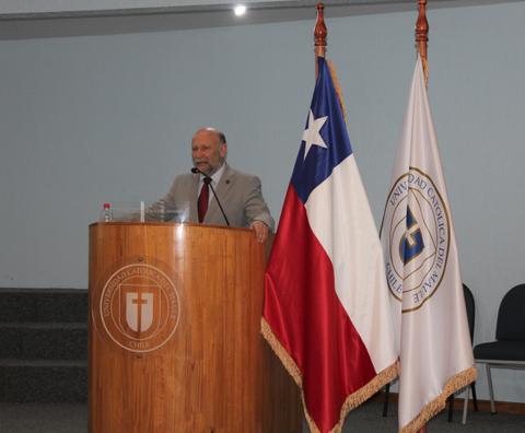 Un hombre de pie frente a un podio de madera con banderas de Chile y de una universidad católica.