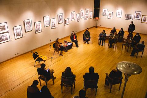 Un grupo de personas sentadas en un salón con fotografías en las paredes, mientras una mujer presenta una actuación musical.