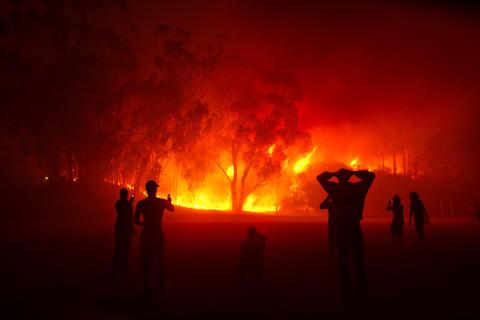 Una escena de un incendio forestal con llamas y humo que rodea a varias personas que observan.