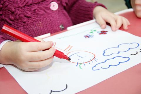 Una niña dibuja con un marcador rojo en una hoja de papel con nubes y flores.