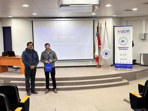 Dos hombres se encuentran en un auditorio junto a una pantalla y un banner institucional.