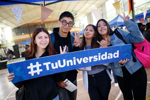 Un grupo de cuatro jóvenes sonríen y posan juntos sosteniendo un letrero que dice #TuUniversidad.
