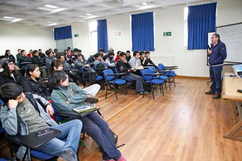 Una clase en un aula con alumnos prestando atención al profesor que está de pie frente a ellos.
