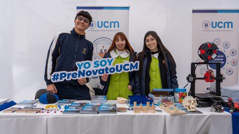 Tres jóvenes posan en un evento con un cartel que dice 'YO SOY #PreNovatoUCM' junto a una mesa con materiales educativos.