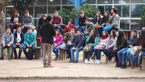 Un grupo de estudiantes sentados en bancas mientras uno de ellos habla frente a ellos.